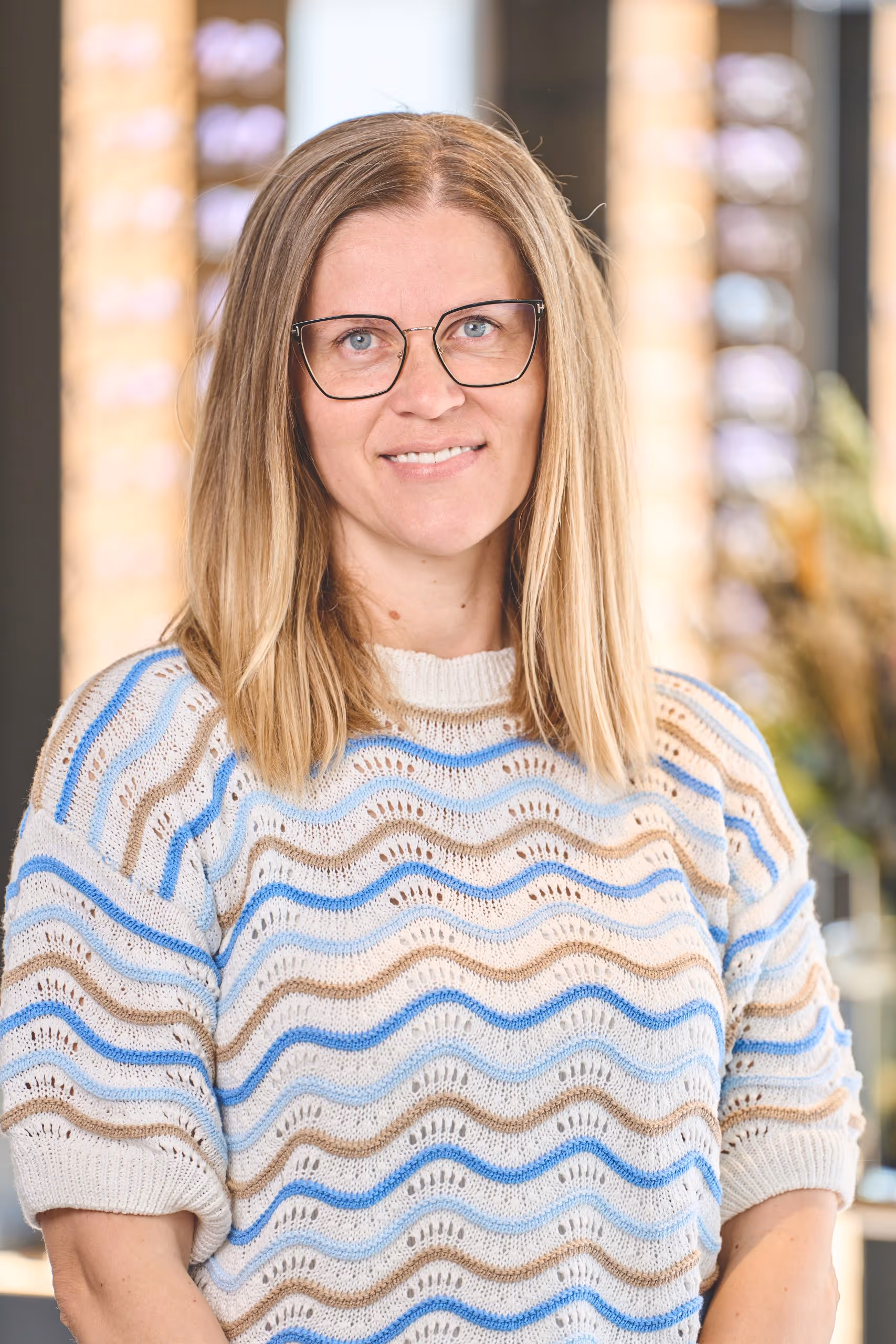 Smiling woman with blonde hair and glasses wearing a knitted sweater with blue and beige wavy stripes.