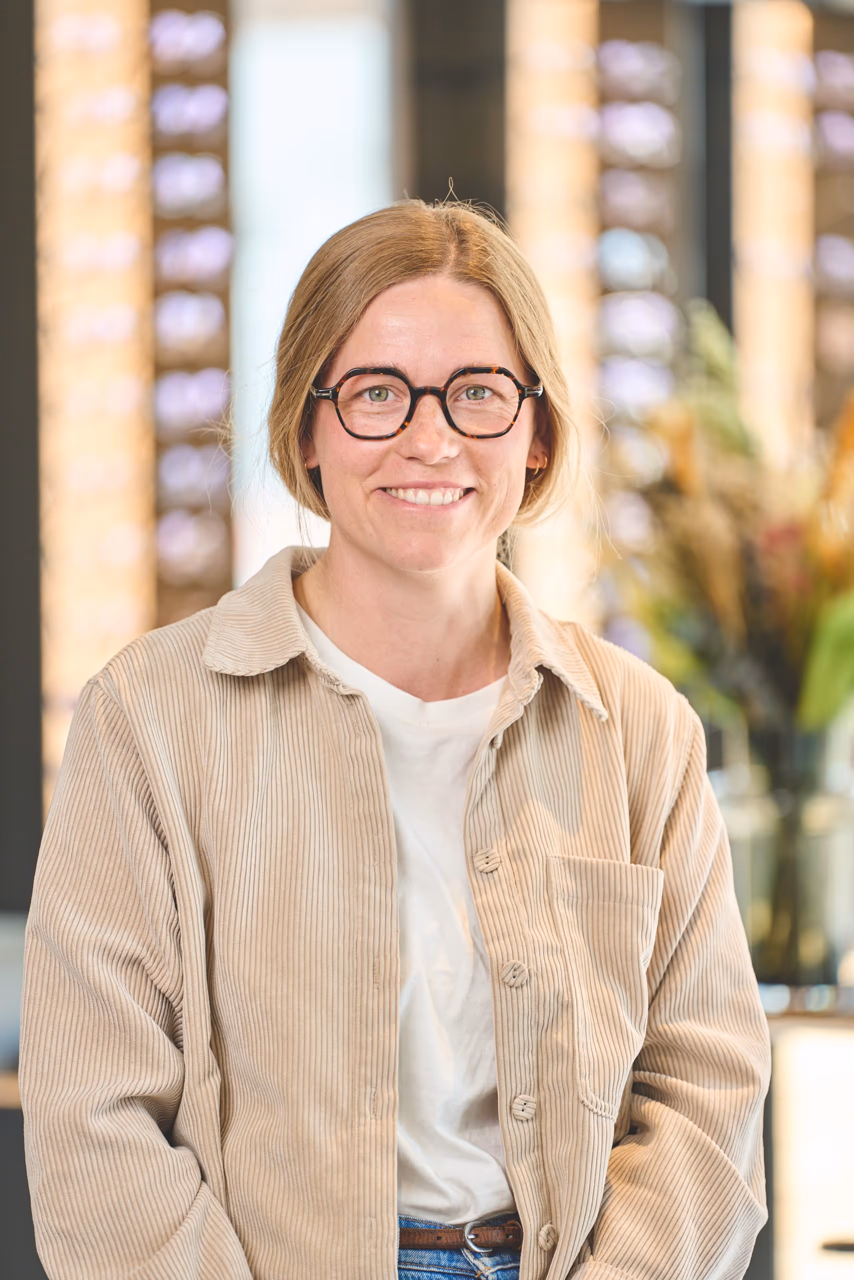 Smiling woman with light brown hair, glasses, beige corduroy jacket, and white T-shirt in a bright indoor setting.