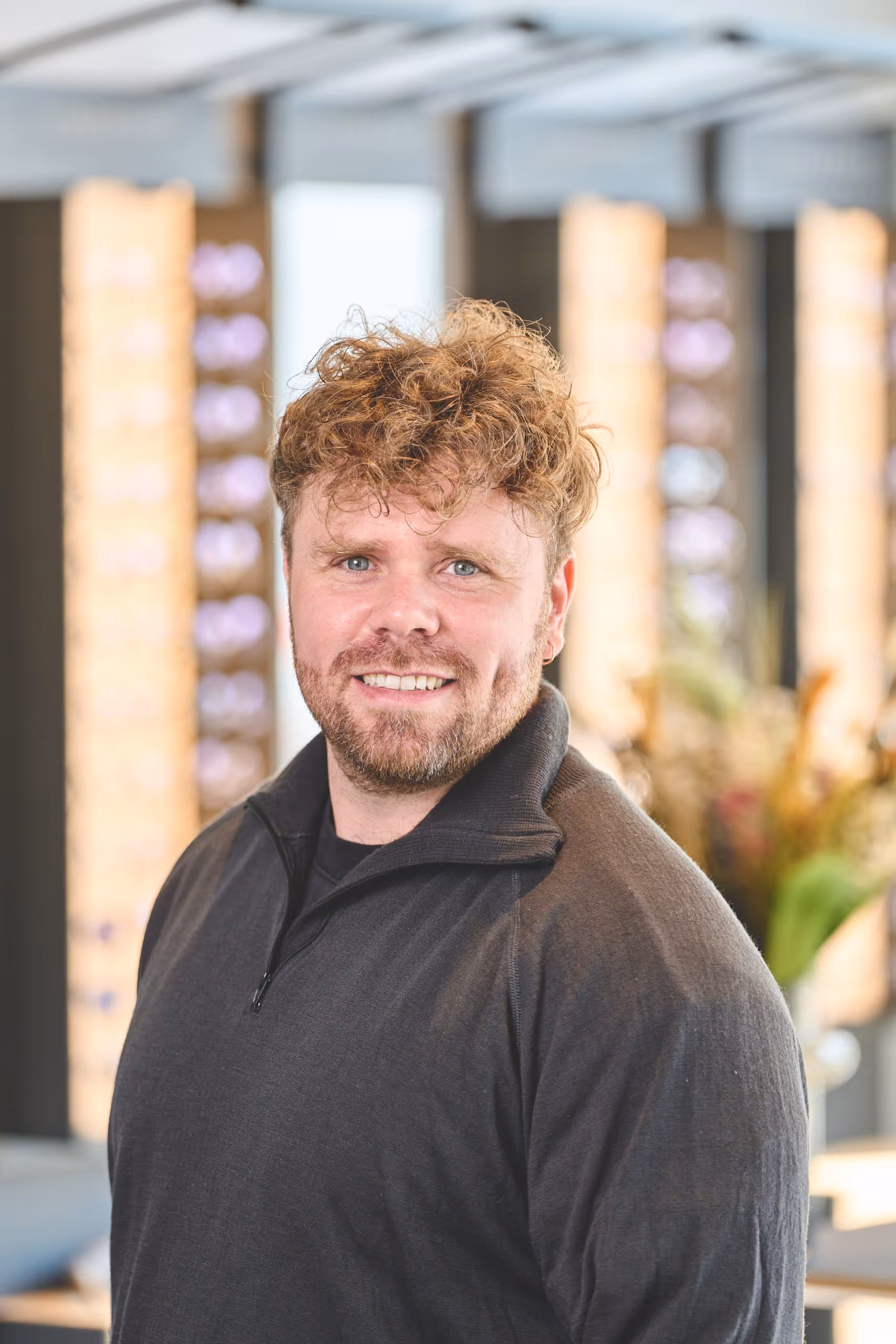 Smiling man with curly hair and beard wearing a black quarter-zip sweater in a bright indoor space.