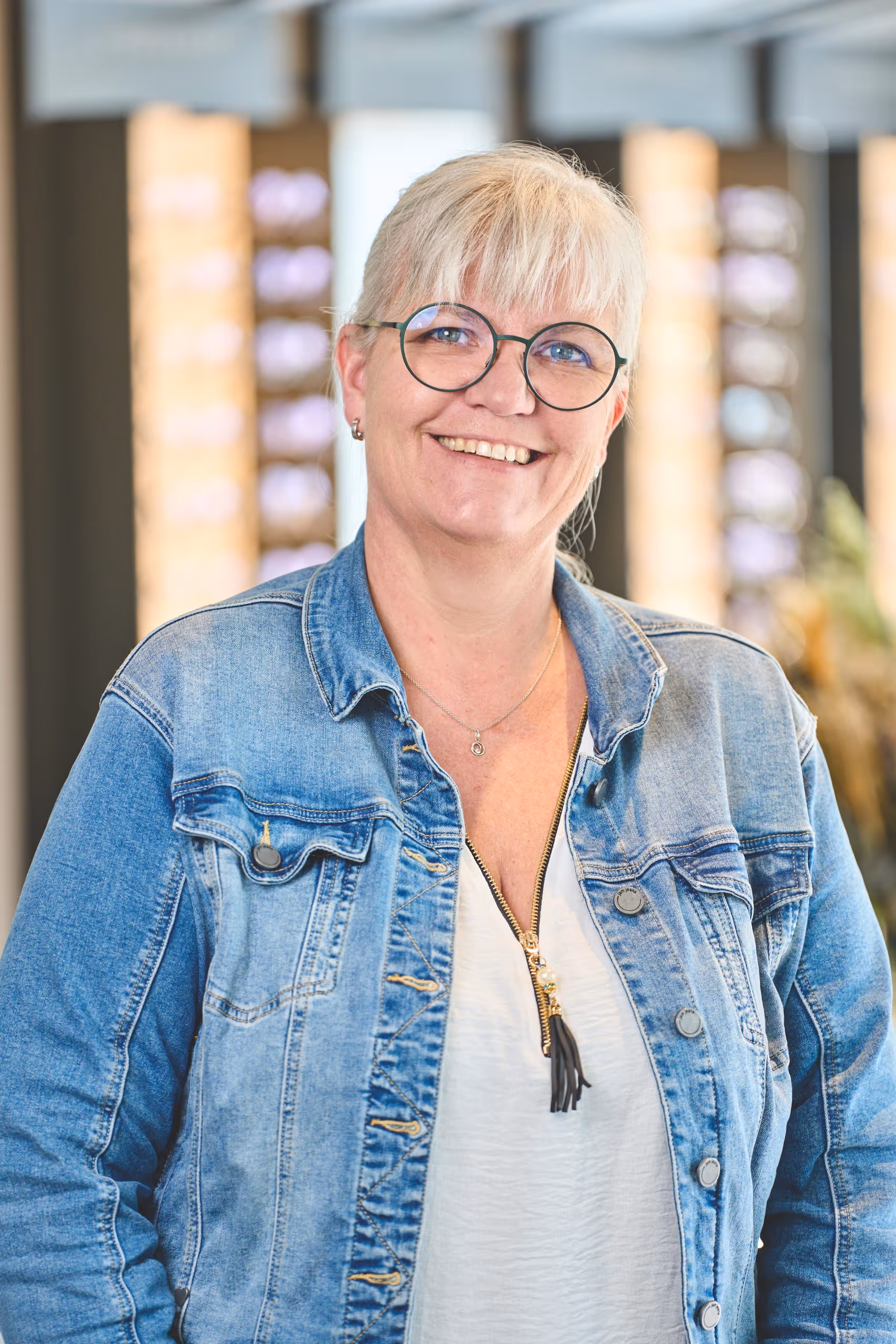Smiling woman with short blonde hair, round glasses, wearing a blue denim jacket and white shirt indoors.