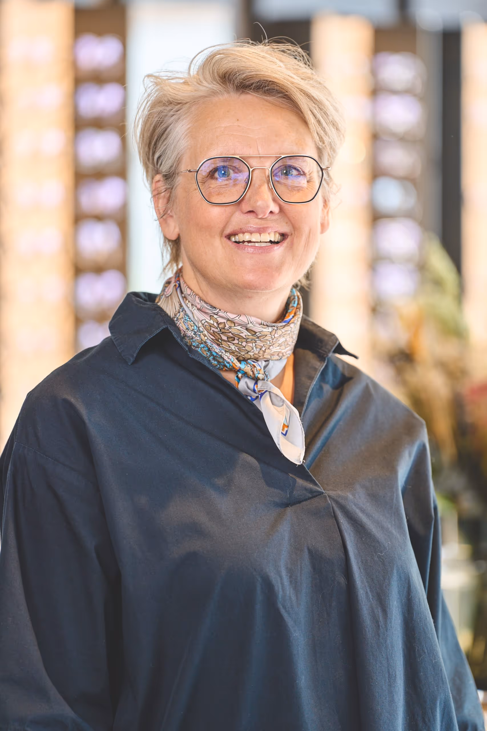 Smiling middle-aged woman with short blonde hair, wearing glasses, a dark shirt, and a patterned scarf around her neck.
