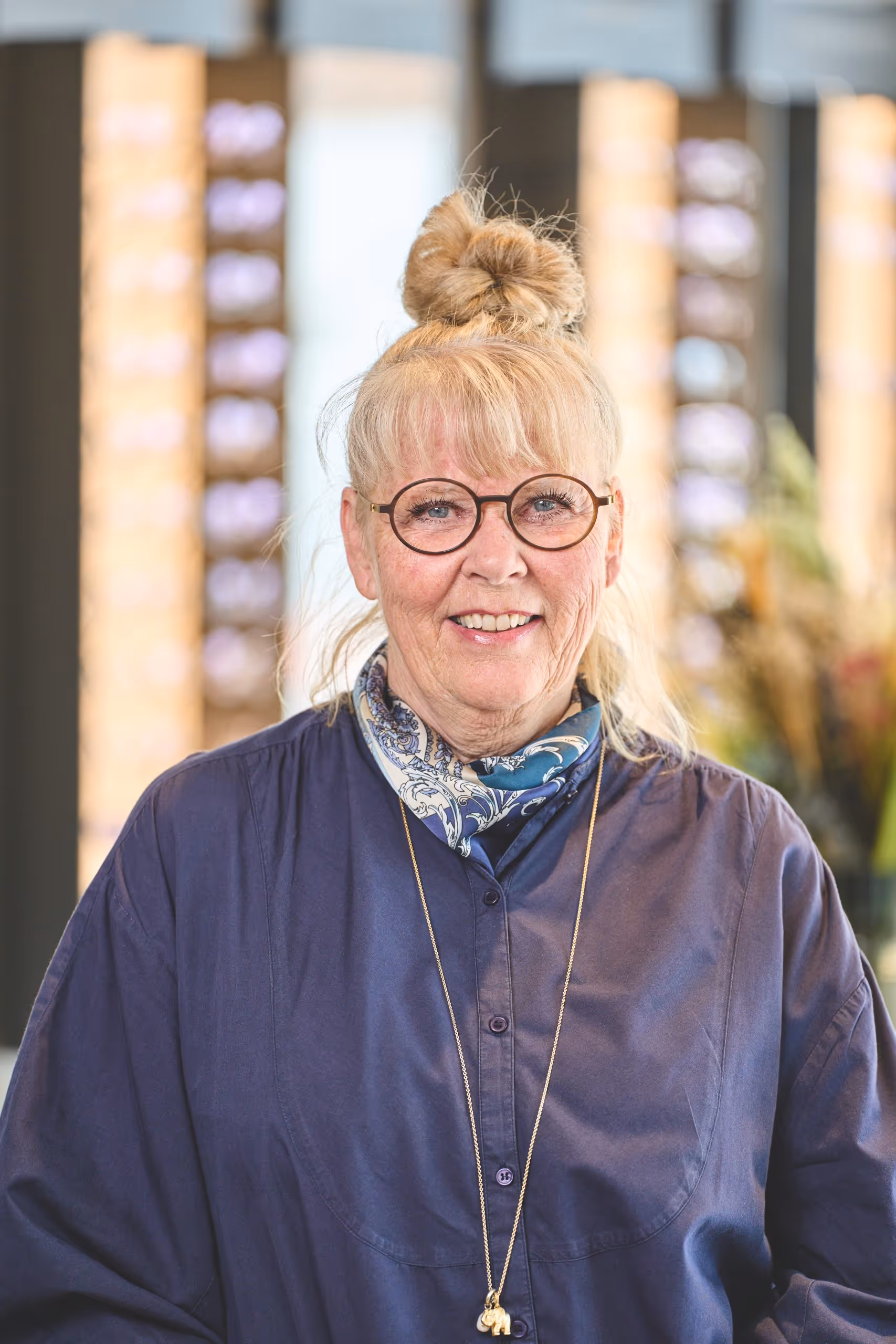 Smiling older woman with blonde hair in a top bun, wearing round glasses, a blue patterned scarf, navy shirt, and an elephant pendant necklace.