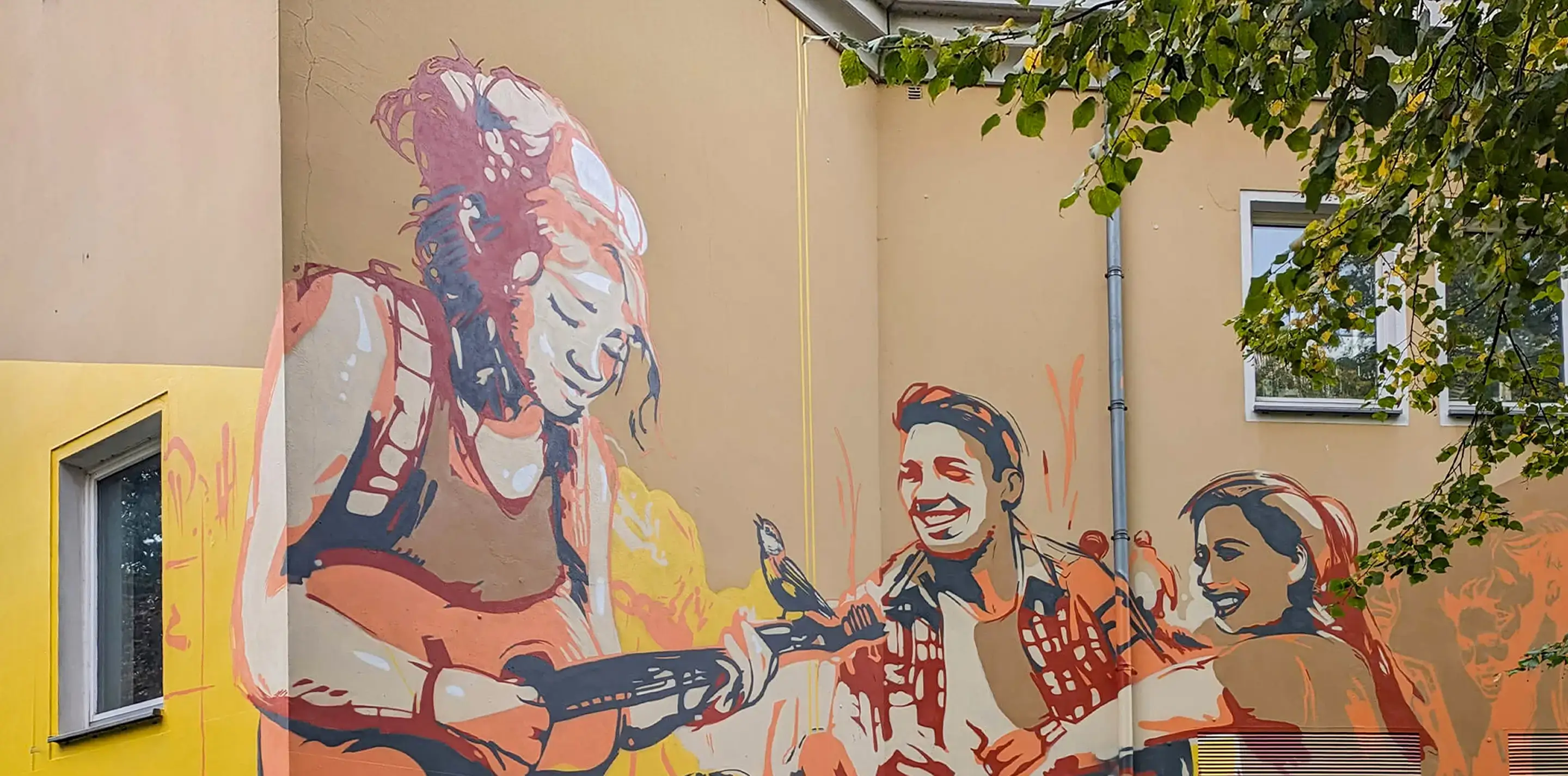 Großflächiges Graffiti am Verwaltungsgebäude des Studierendenwerks Osnabrück zeigt drei sitzende Personen, eine mit Gitarre, in warmen Gelb- und Orangetönen.