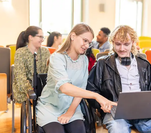 Junge Frau im Rollstuhl zeigt einem jungen Mann mit Kopfhörern Informationen auf einem Laptop in einem Besprechungsraum.