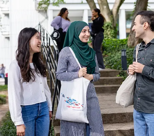 Drei Studierende unterhalten sich draußen auf einem Campusgelände vor einem Treppenaufgang, eine der beiden Frauen trägt ein grünes Kopftuch.