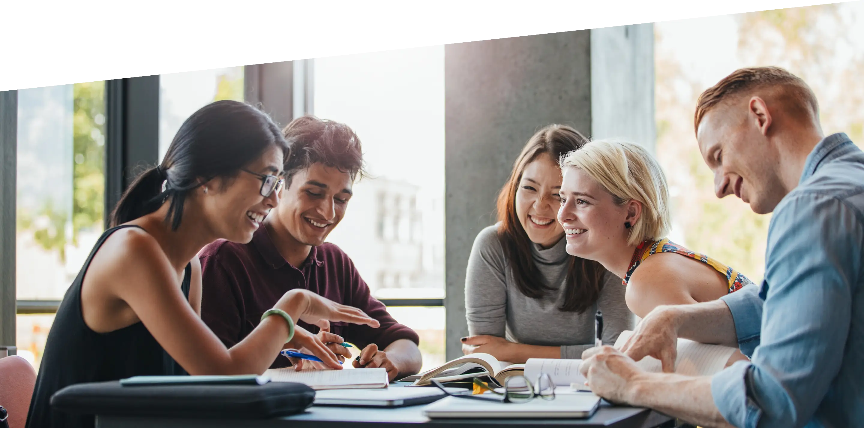 Eine Gruppe von fünf jungen Erwachsenen sitzt an einem Tisch, lacht und lernt gemeinsam mit Büchern und Notizblöcken.