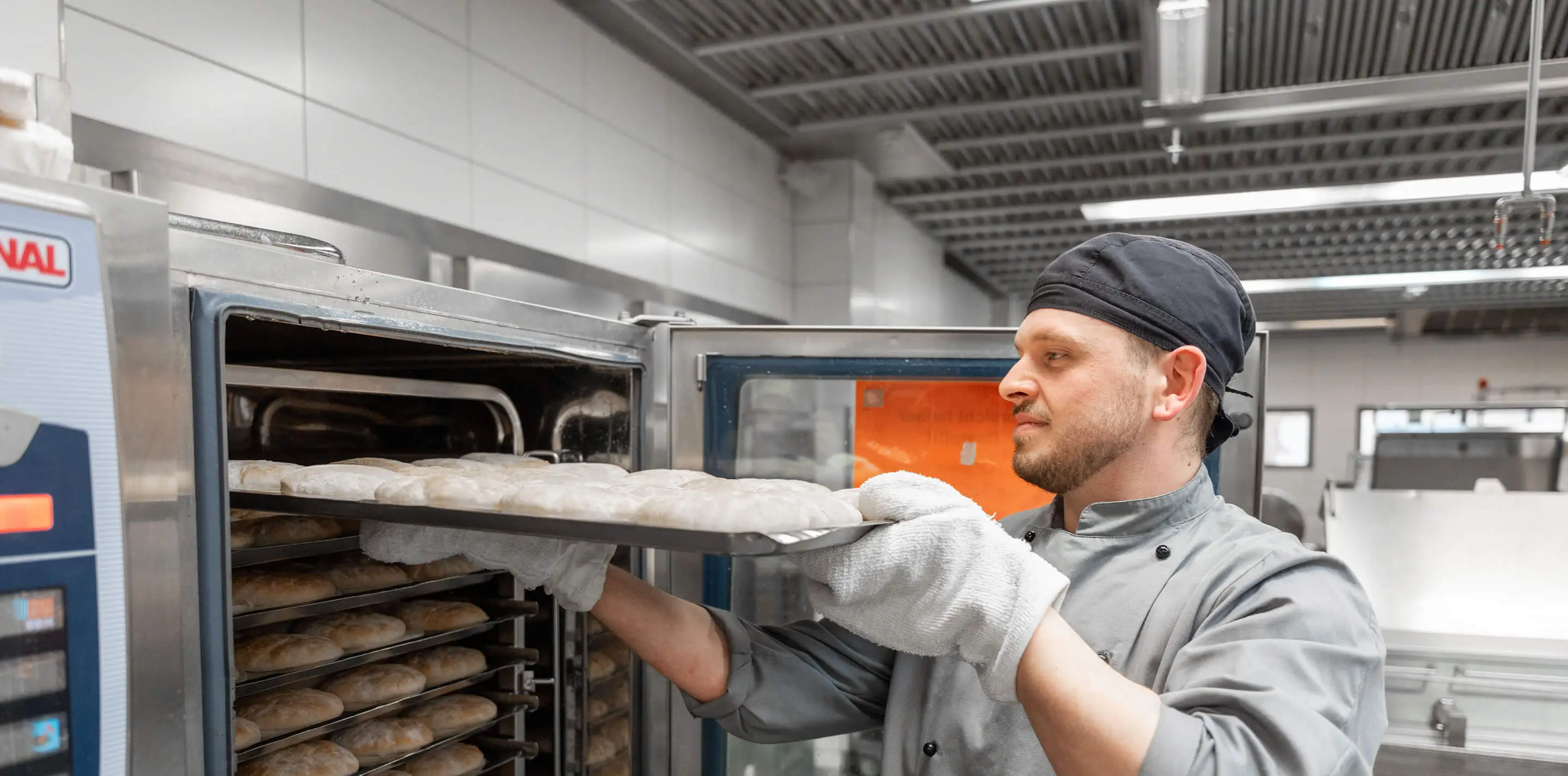 Ein Bäcker entnimmt ein Backblech mit rohen Brötchen aus einem großen Kühlschrank in der Hochschulküche.