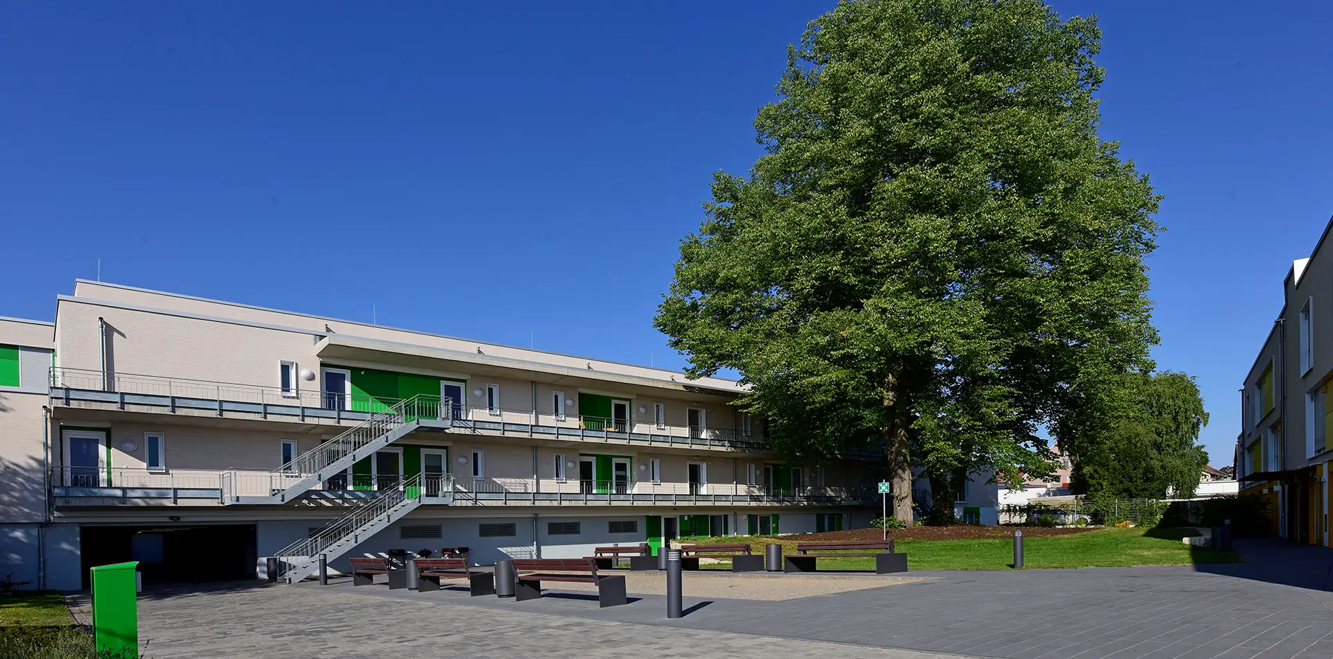 Moderner Wohnkomplex mit weißen Gebäuden, grünen Türen, Außenstiegen und großen Bänken in einer kleinen Grünanlage mit Wiese.