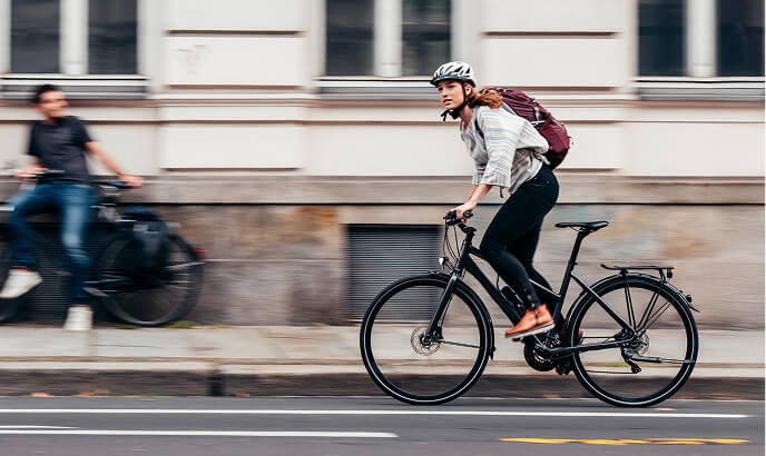 Eine Frau mit Helm und Rucksack fährt in der Stadt mit einem Fahrrad auf der Straße vor einem Gebäude mit großen Fenstern.