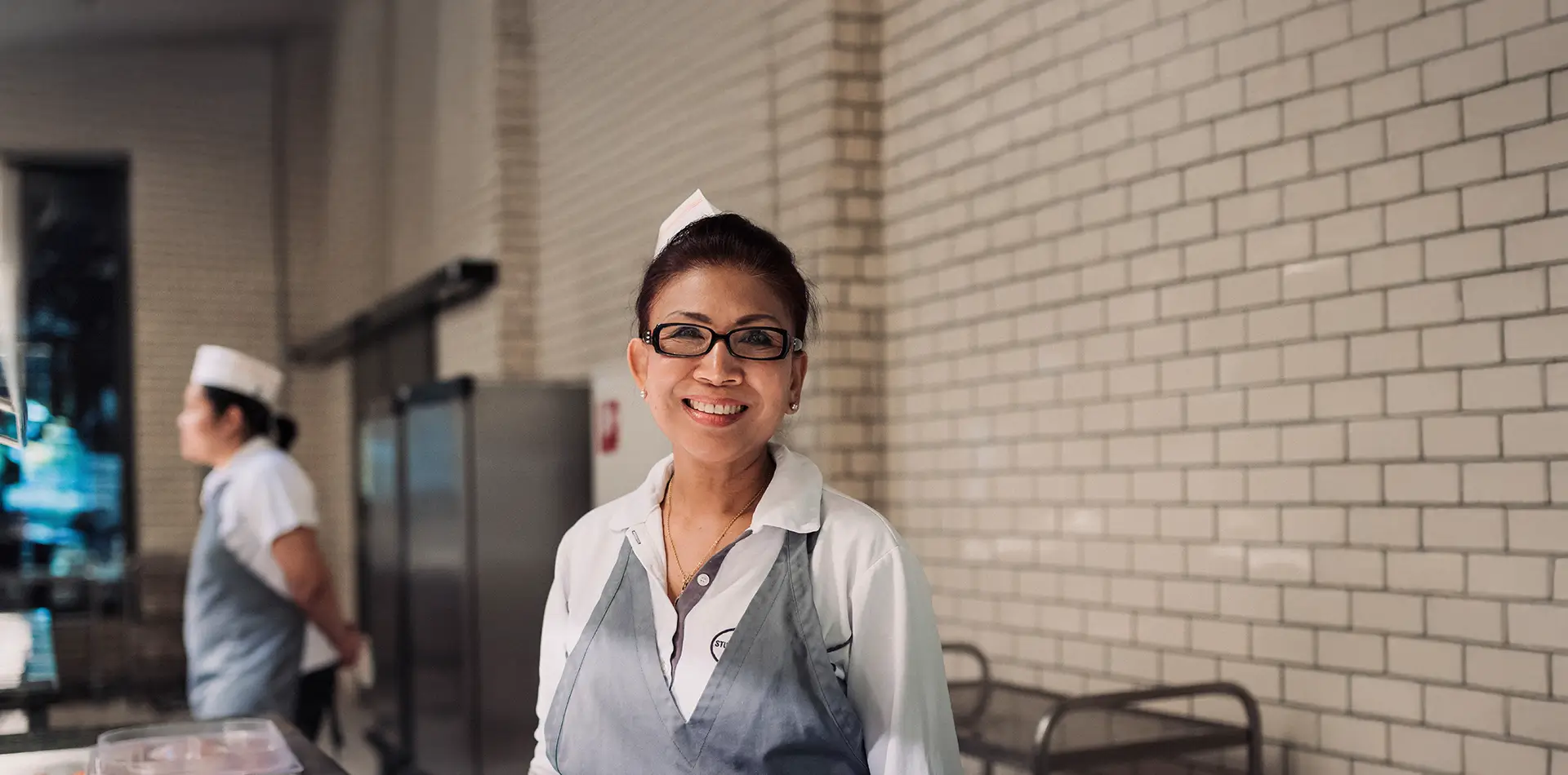 Lächelnde Frau in Kellneruniform mit Brille steht vor einer Buffetausgabe in einer Mensa und blickt in die Kamera.