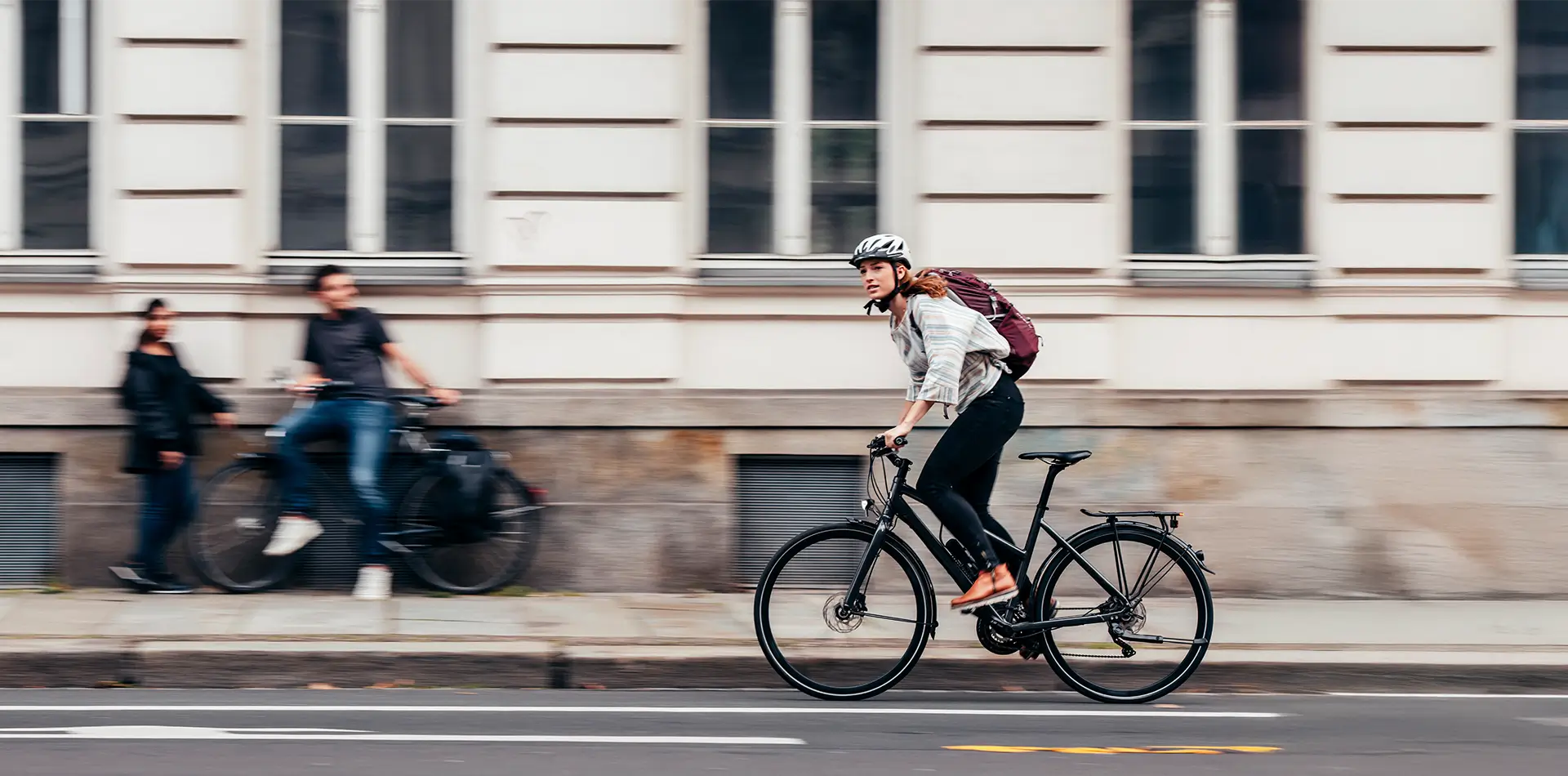 Eine Frau mit Helm und Rucksack fährt in der Stadt mit einem Fahrrad auf der Straße vor einem Gebäude mit großen Fenstern.