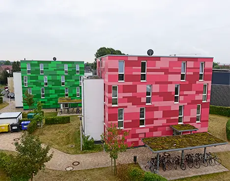 Zwei mehrstöckige Wohngebäude mit bunten Fassaden in verschiedenen Grüntönen und Rottönen umgeben von gepflegten Grünflächen und Fahrradständern mit einer Überdachung.