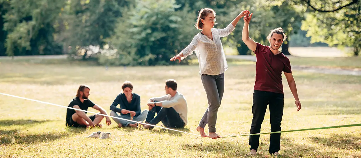 Eine Frau balanciert barfuß auf einem Slackline-Band im Park, während ein Mann ihre Hand hält. Drei weitere junge Personen sitzen im Hintergrund auf der Wiese.