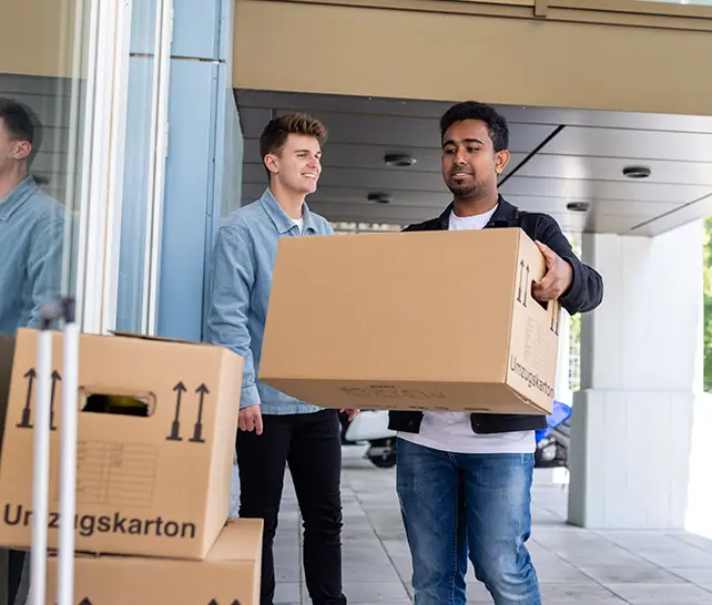 Zwei junge Männer tragen Umzugskartons vor einem modernen Gebäude.