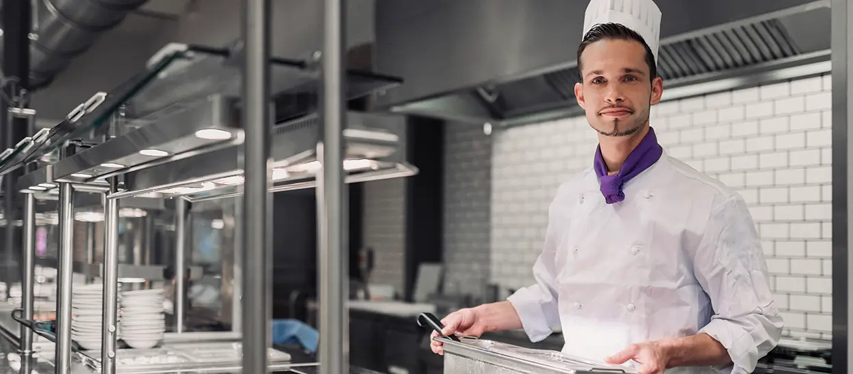 Koch in weißer Uniform mit lila Halstuch hält ein Tablett in einer professionellen Küchenumgebung und blickt in die Kamera.