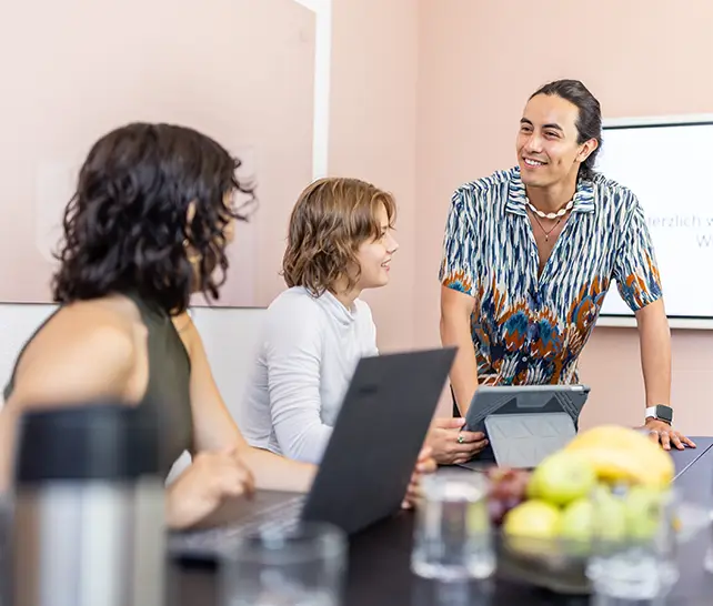 Drei Personen in einem Meetingraum unterhalten sich, zwei sitzen mit Laptops am Tisch, eine steht und lächelt.