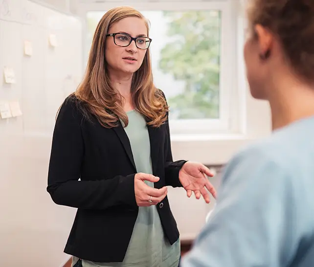 Frau mit Brille und schwarzem Blazer erklärt etwas vor einem Whiteboard mit Haftnotizen in einem Büro.