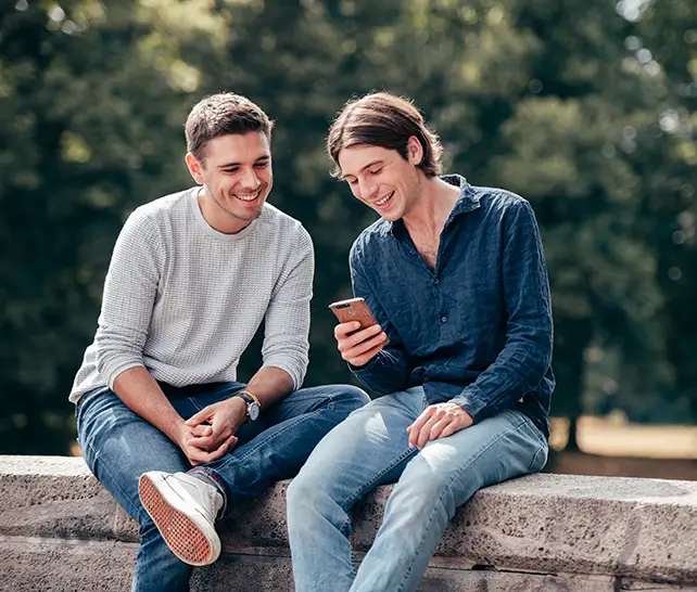 Zwei junge Männer sitzen auf einer Steinmauer im Freien und schauen lachend auf ein Smartphone.
