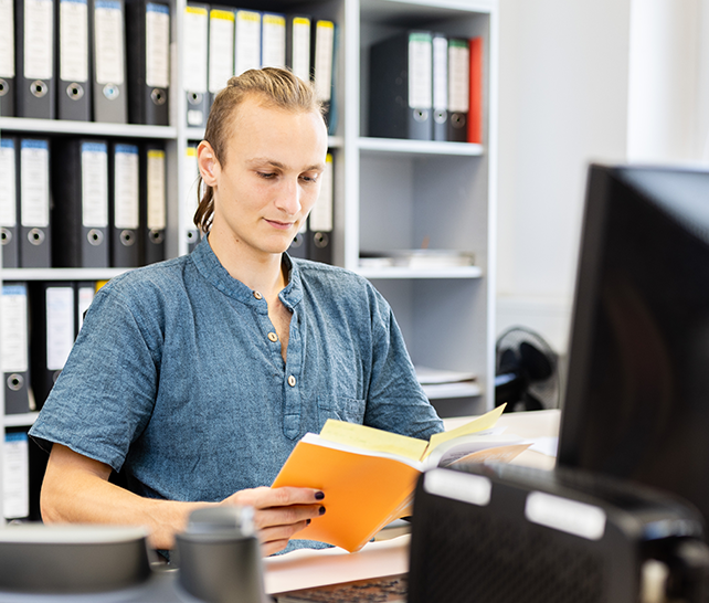 Junger Mann sitzt am Schreibtisch in einem Büro und liest in einem Handbuch.