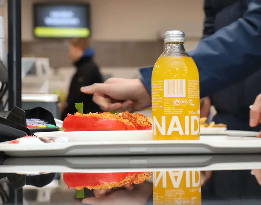 Nahaufnahme eines Tabletts mit einer gefüllten roten Paprika und einer Flasche Orangenlimonade an der Kasse in einer Mensa.