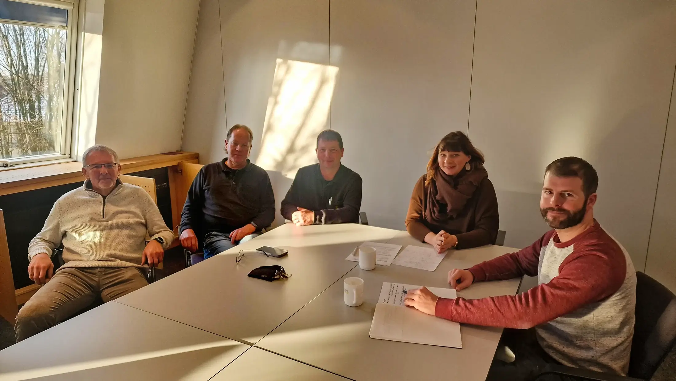 Fünf Personen sitzen um einen Besprechungstisch in einem hellen Raum mit Fenster und Unterlagen.