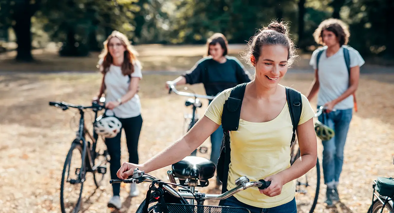 Vier junge Menschen schieben ihre Fahrräder auf einem Weg in einem Park mit Bäumen im Hintergrund.