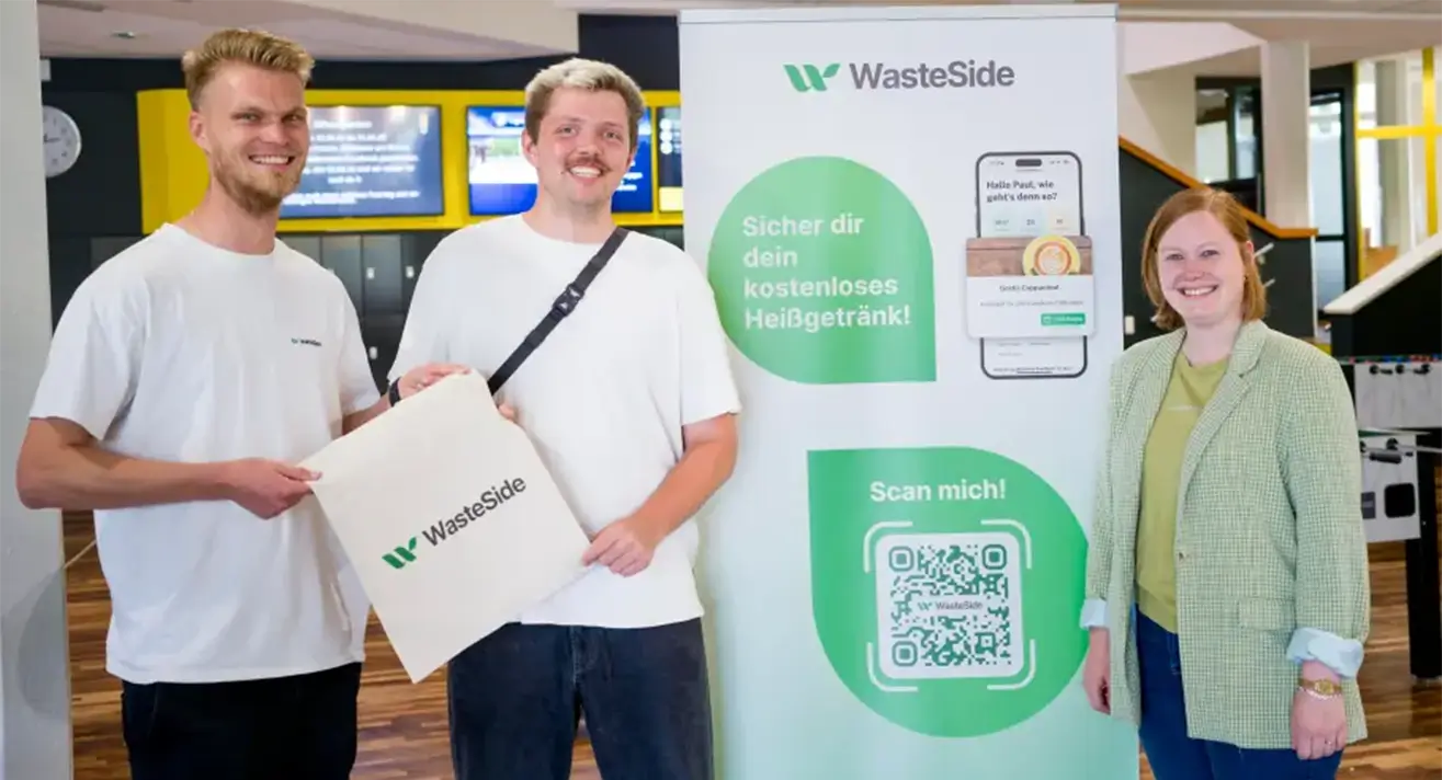 Drei Personen stehen vor einem Rollup mit der Überschrift WasteSide und einem Handy-Mockup. Zwei halten eine WasteSide-Tasche.