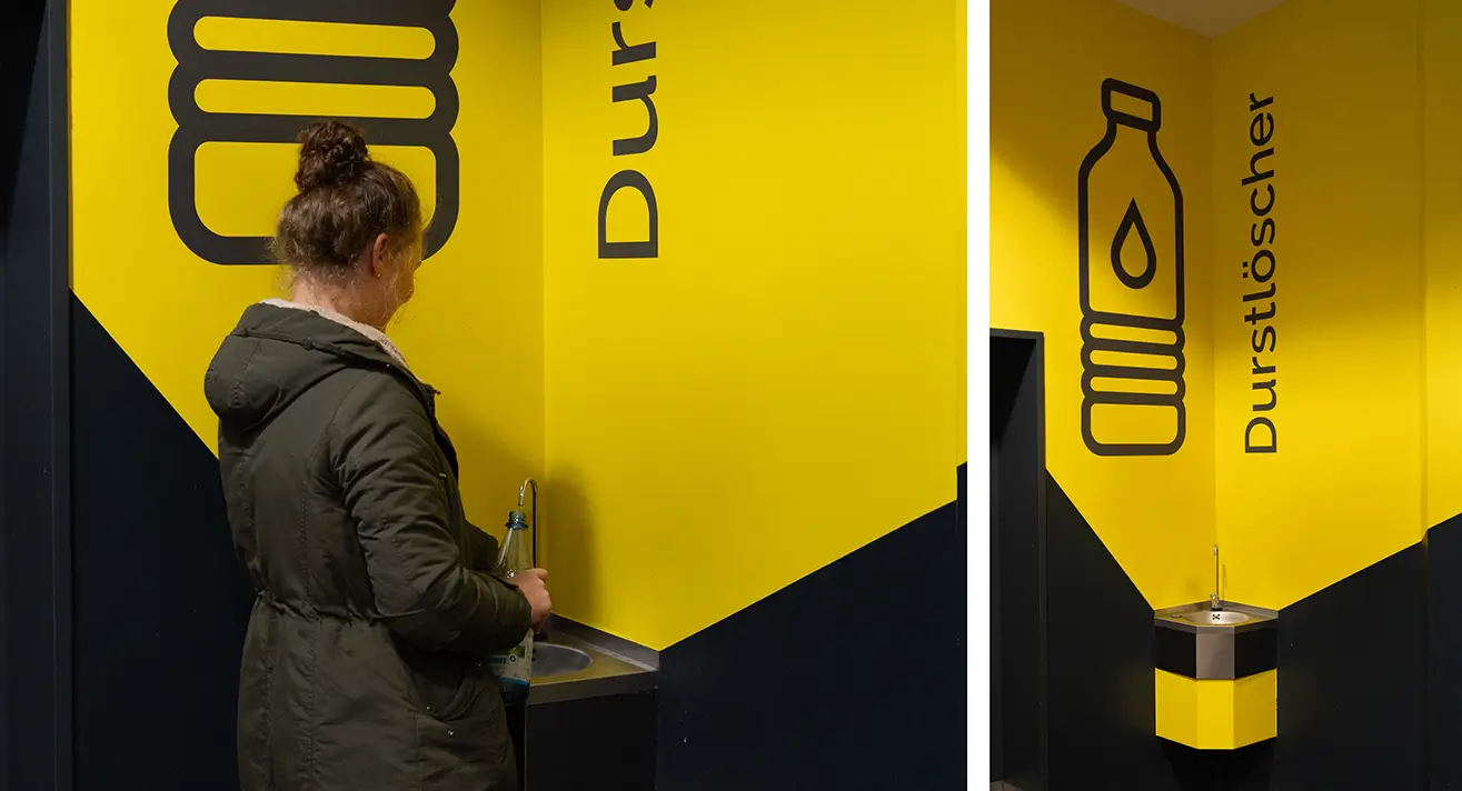 Eine Frau füllt eine Flasche an einem Wasserspender vor gelb-schwarzer Wand mit Schrift auf.