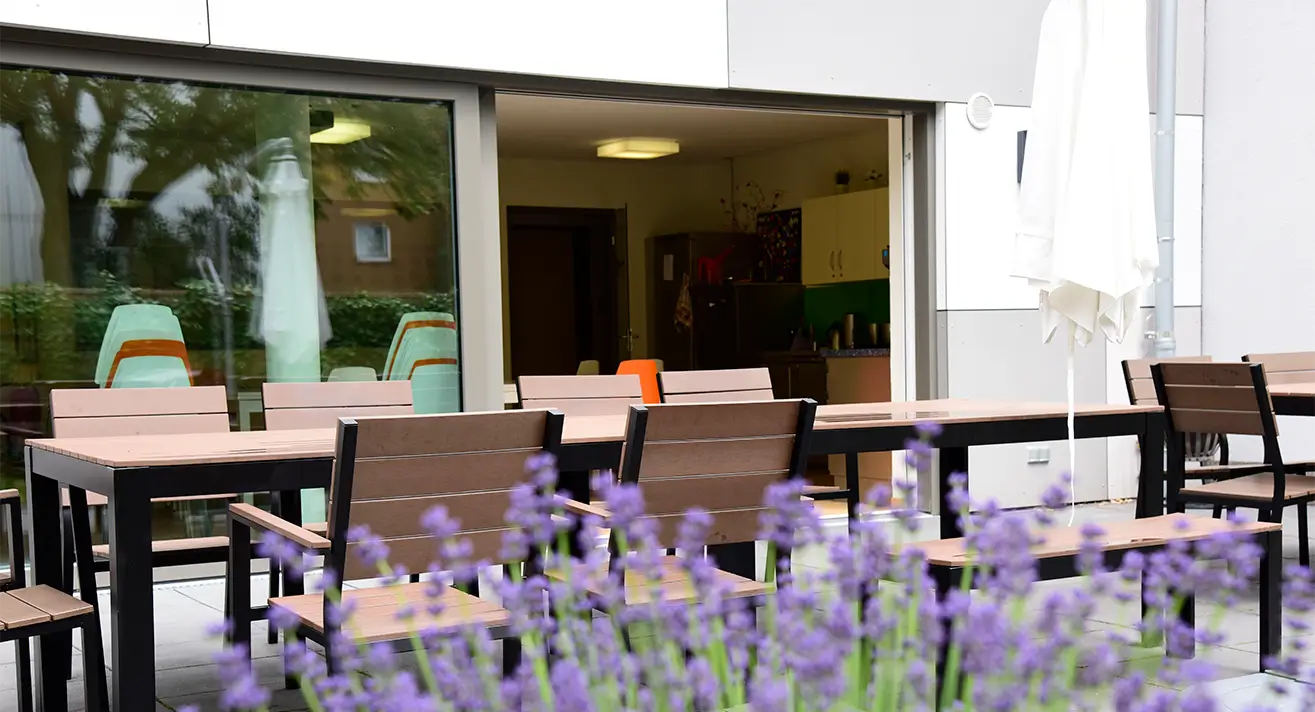 Terrasse mit mehreren braunen Holztischen und Stühlen vor einem Gebäude, im Vordergrund violette Blüten, im Hintergrund ein offenes Fenster mit Blick in einen Raum mit grüner Küchenzeile.