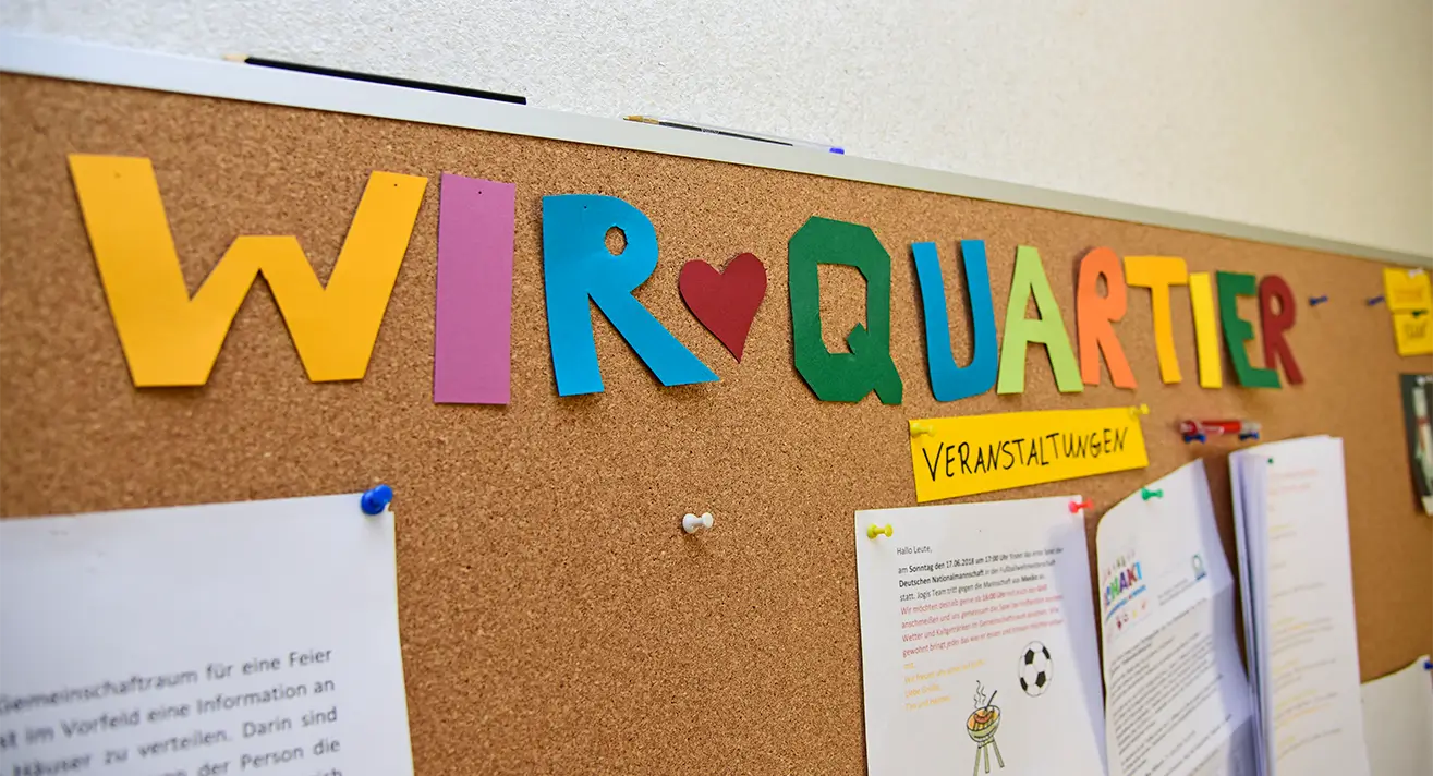 Bunte Buchstaben auf einer Pinnwand bilden den Schriftzug 'WIR ♥ QUARTIER', darunter ein gelber Zettel mit dem Wort 'VERANSTALTUNGEN'.