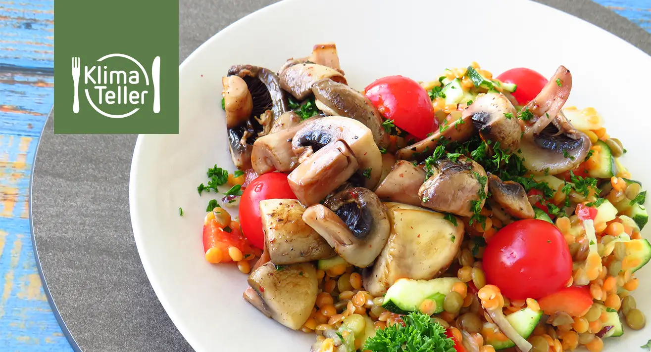 Teller mit Linsensalat, garniert mit Champignons, Kirschtomaten und Petersilie, auf grauem Platzset und blauem Holztisch, links das Logo 'Klima Teller'.