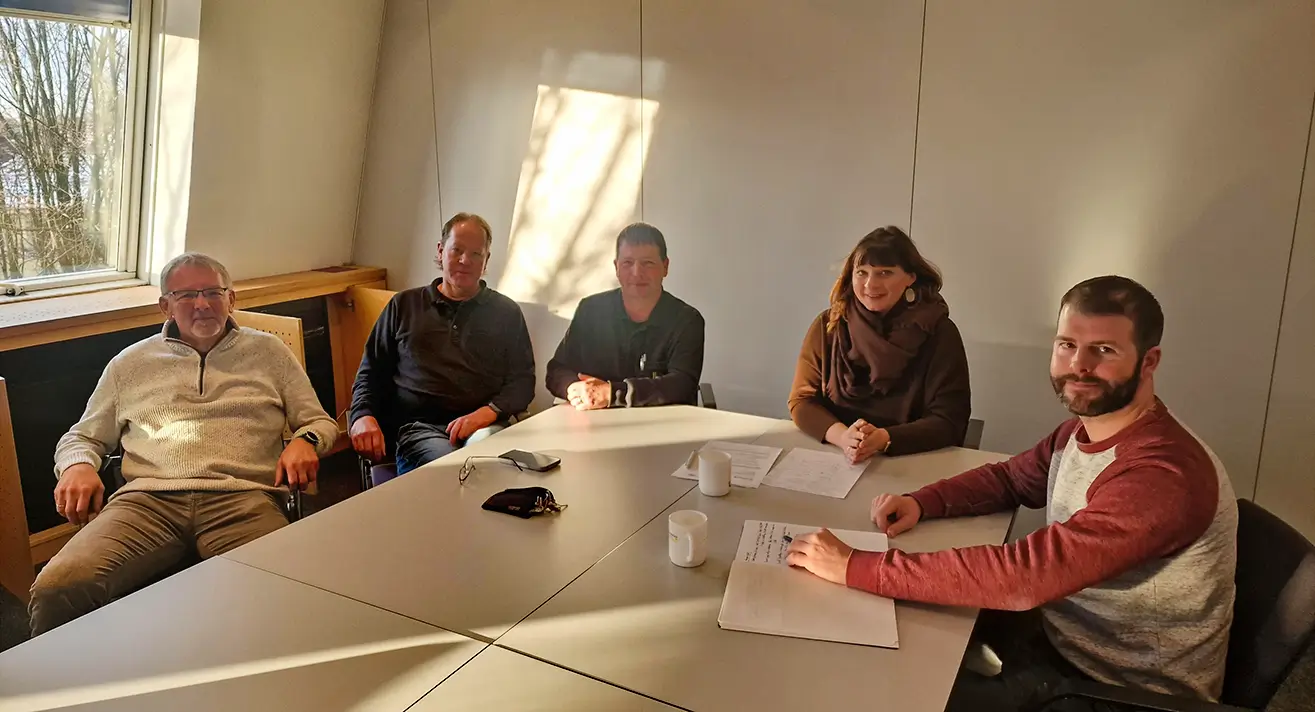 Fünf Personen sitzen um einen Besprechungstisch in einem hellen Raum mit Fenster und Unterlagen.