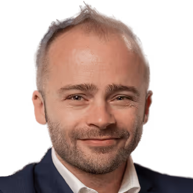 Muž s krátkými vlasy a lehkým strništěm v tmavém obleku a bílé košili se usmívá do kamery.