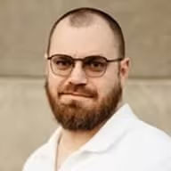 Muž s brýlemi a plnovousem v bílém tričku před neutrálním pozadím.