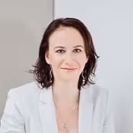 Usměvavá žena s tmavými vlasy v bílém saku na světlém pozadí.