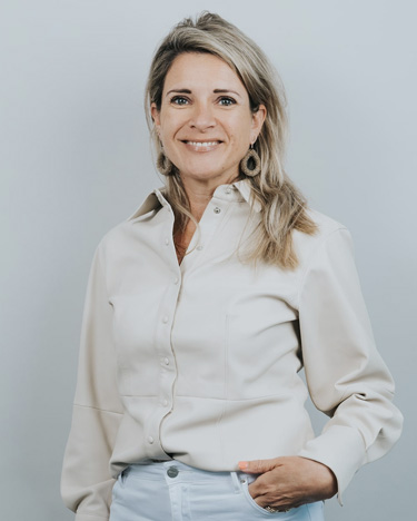 Smiling woman with blonde hair wearing a cream button-up shirt and light pants against a plain gray background.