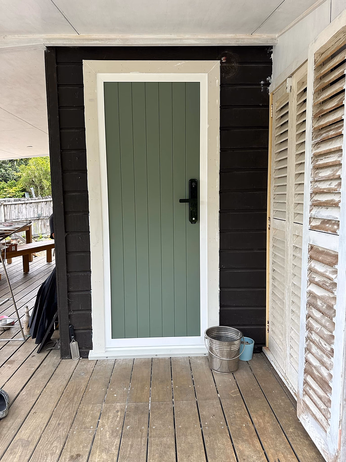 Green vertical panel door with white frame and black handle on a dark wood exterior wall, wooden deck floor in front with metal bucket and small cup to the side.