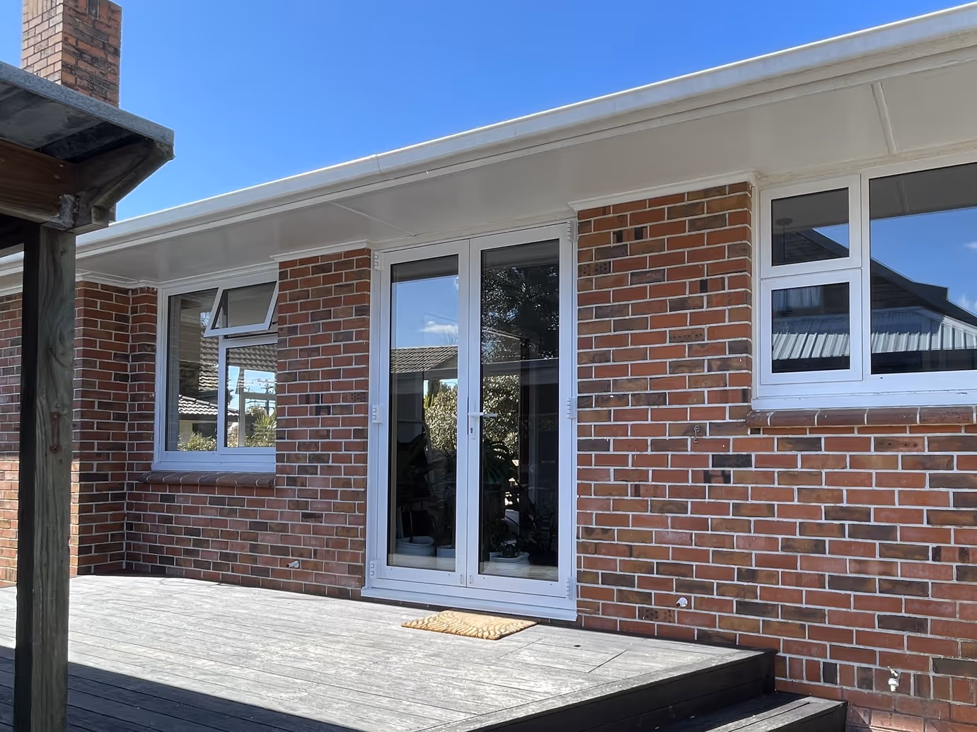 White-framed glass French doors and windows in a red brick house opening onto a wooden deck under a clear blue sky.