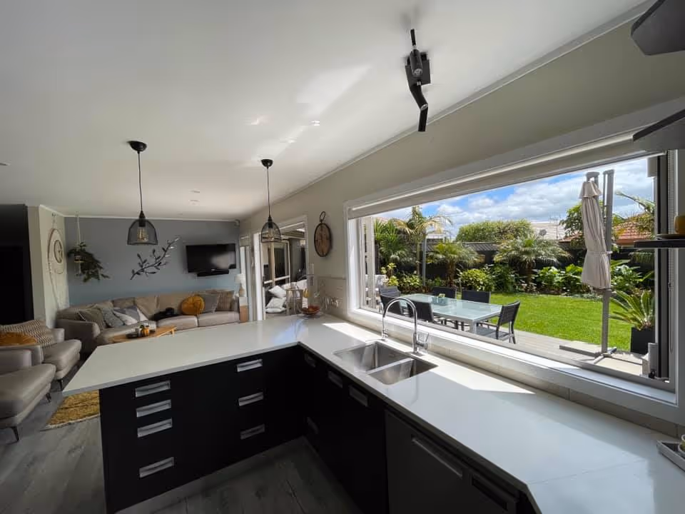 Modern kitchen with white countertops and black cabinets overlooking a garden through a large foldable window, next to a living room with a beige sofa and TV.