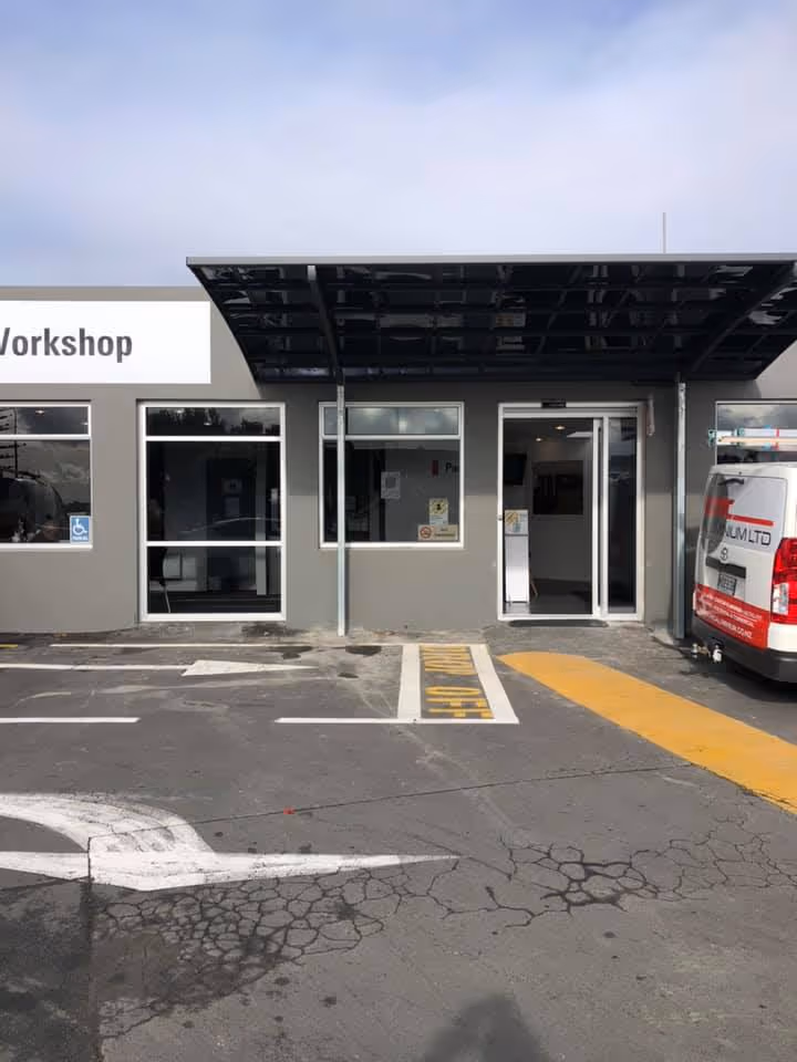 Grey workshop building with a black canopy over sliding glass door entrance and adjacent parking spaces, including a disabled spot and a white van parked on the right.