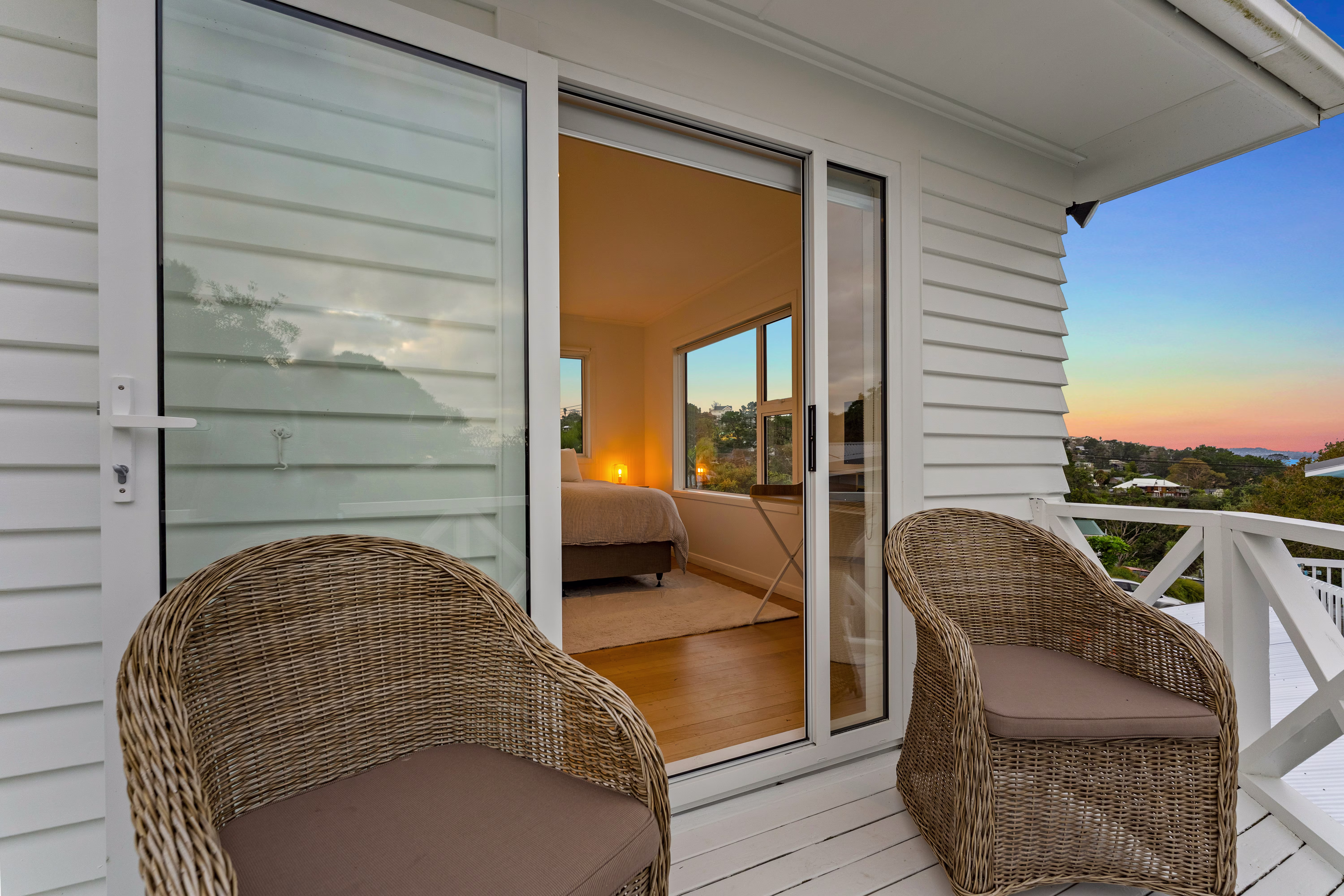 Two wicker chairs with cushions on a white wooden balcony overlooking a sunset view, with a bedroom visible through sliding glass doors.