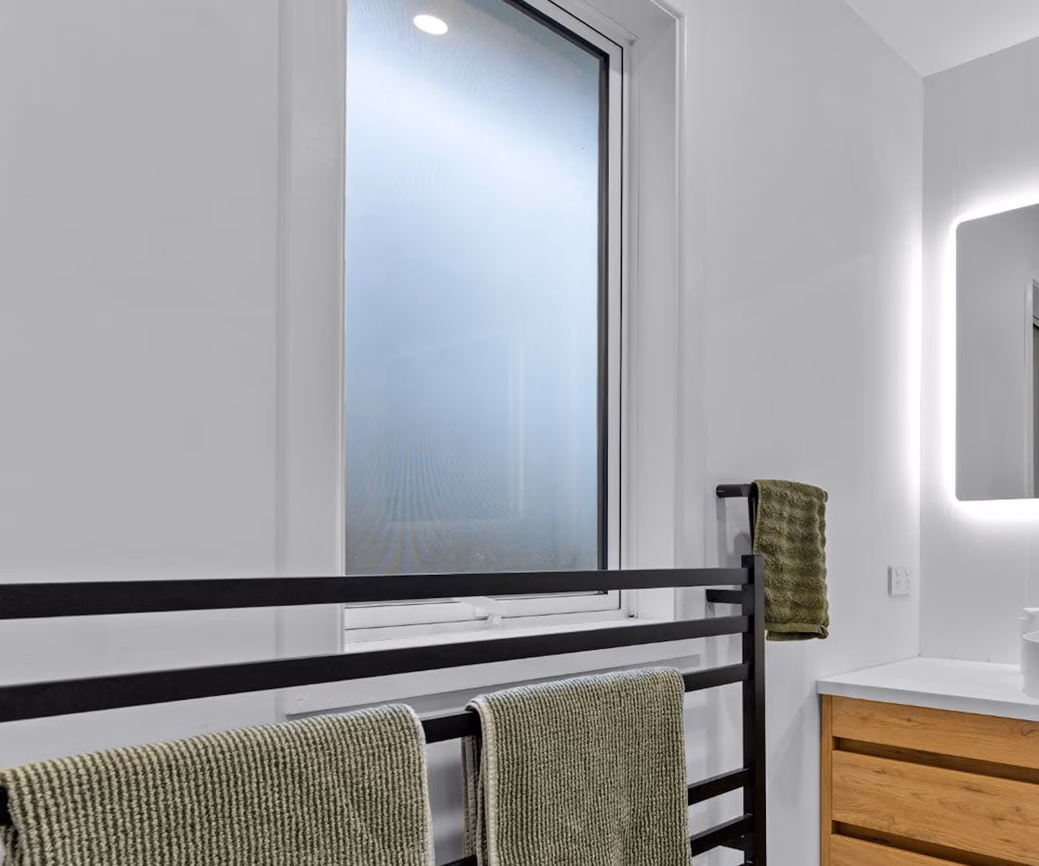 Modern bathroom with frosted window, black towel rack holding green towels, and a wooden vanity with illuminated mirror.