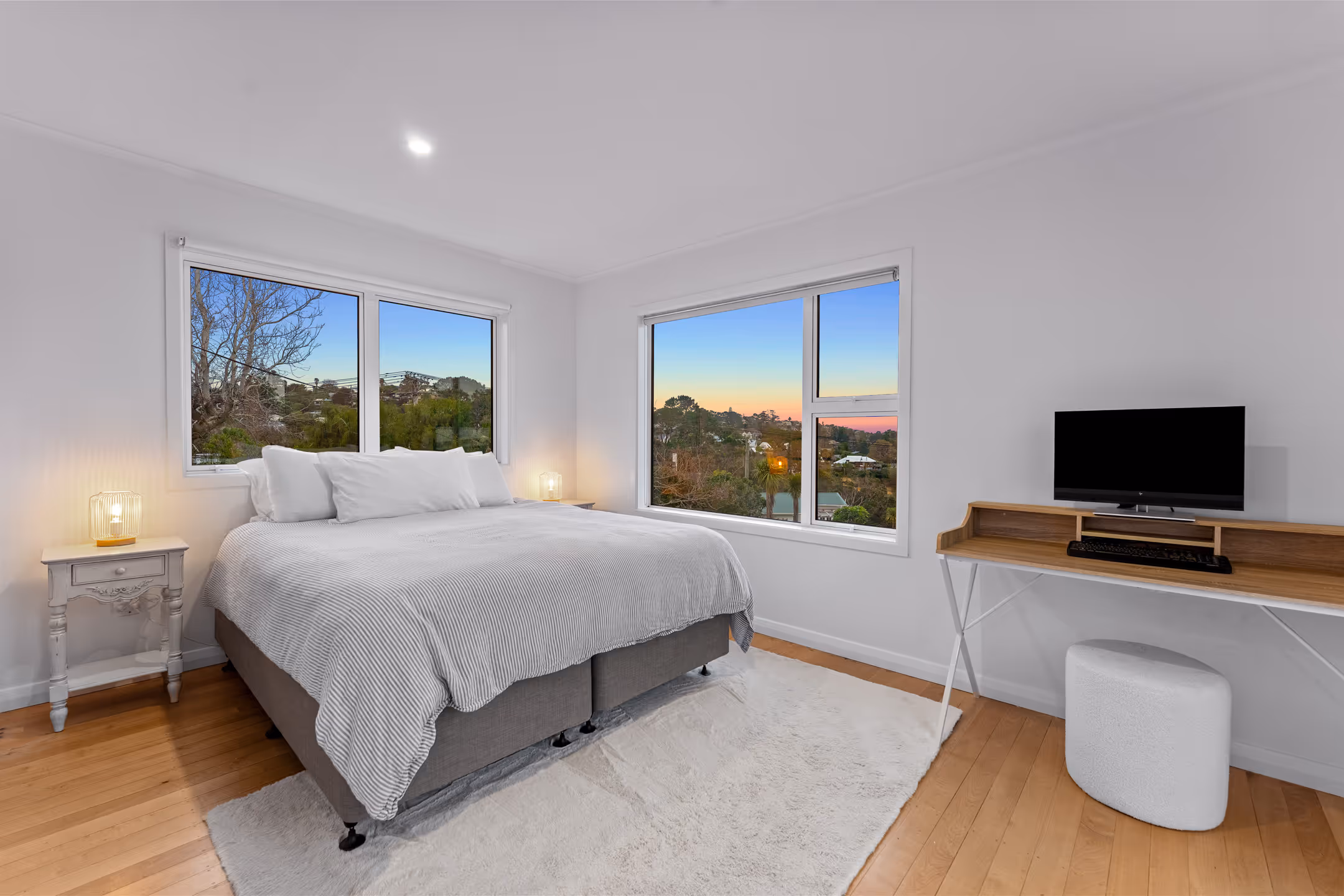 Minimalist bedroom with large bed, two windows showing trees and sunset, wooden floor, white rug, and desk with computer.