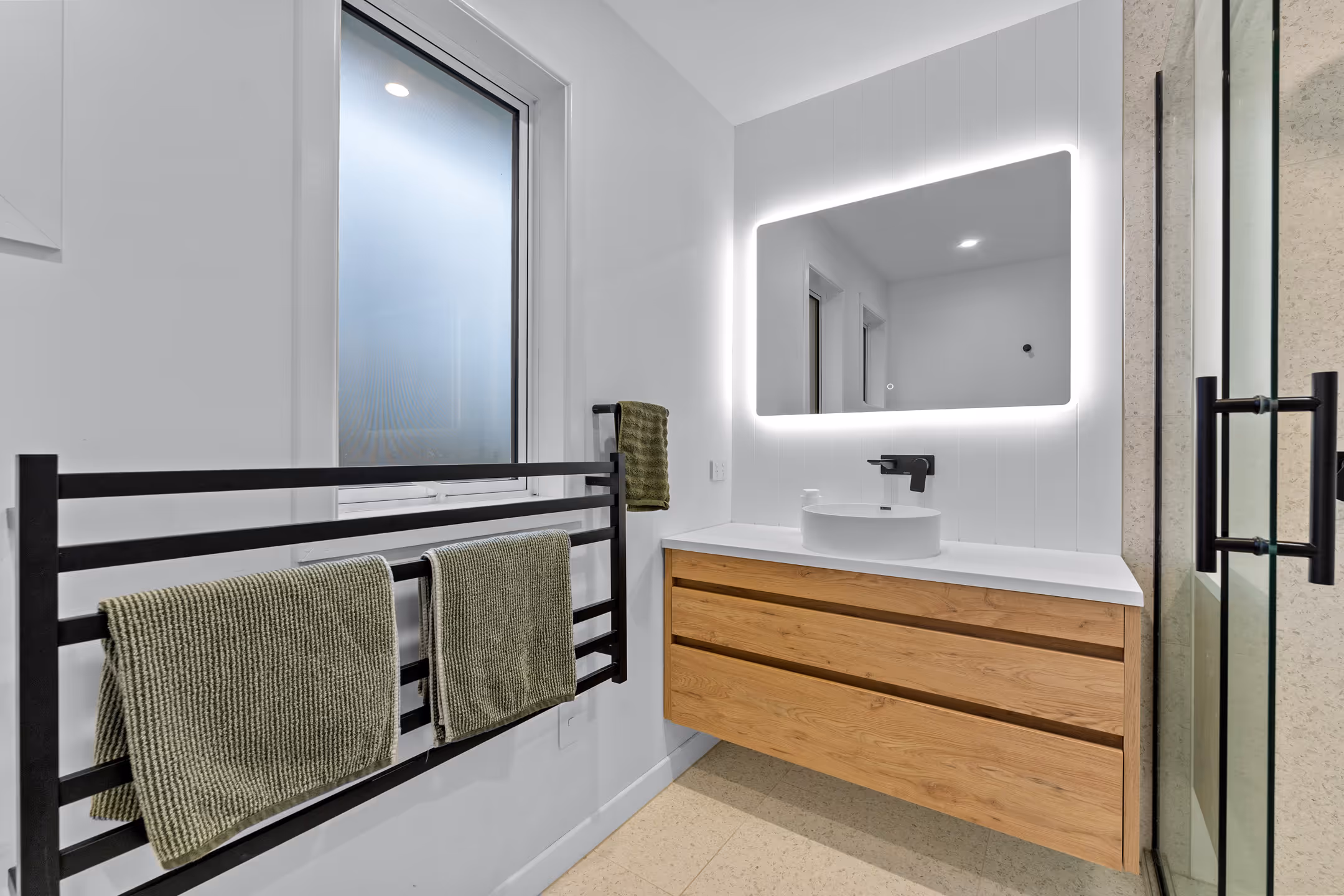 Modern bathroom with wooden vanity, round white sink, illuminated rectangular mirror, towel rack with green towels, and frosted window.