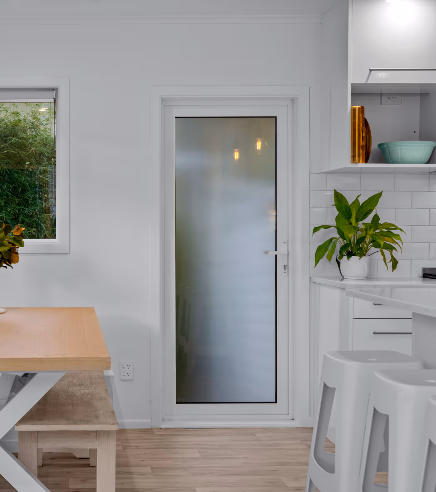 Modern kitchen and dining area with a frosted glass door, wooden table with bench seating, white bar stools, and green plants.