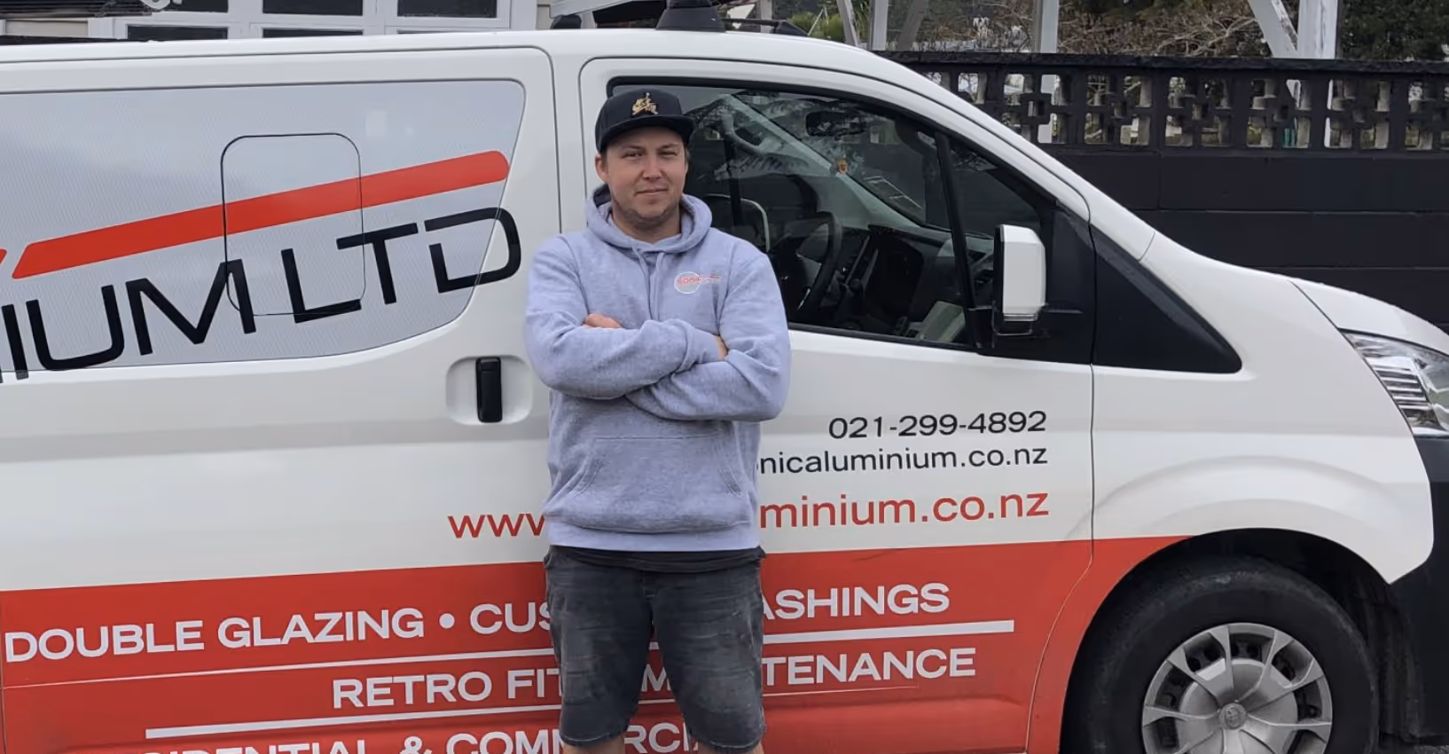 Man wearing a grey hoodie and black cap standing with arms crossed in front of a white and red company van advertising aluminium services.