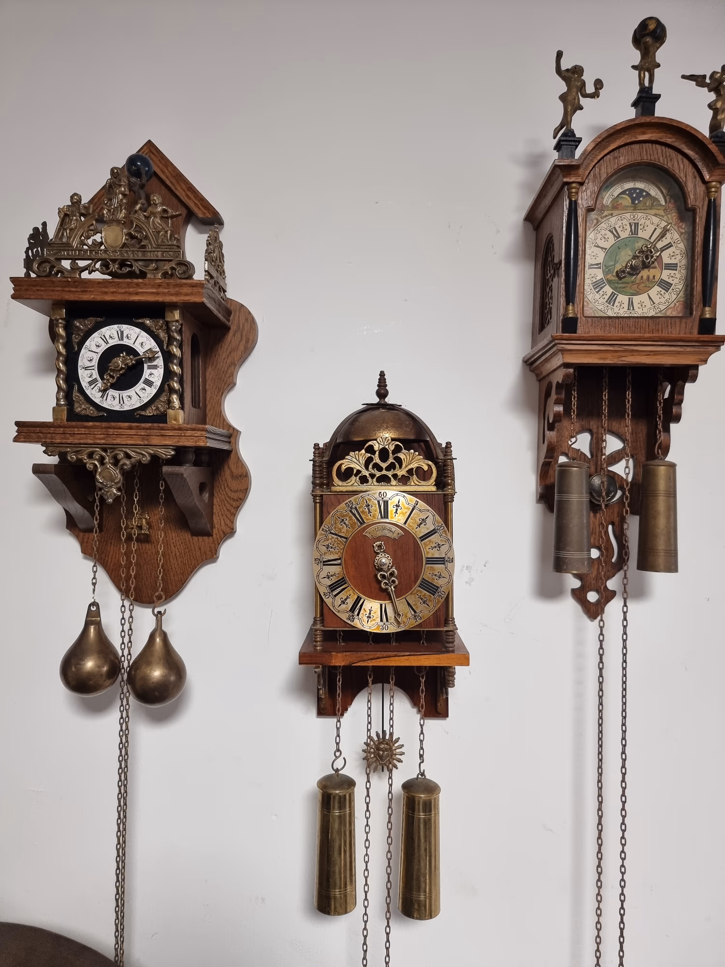 שלוש שעוני קיר עתיקים עם משקולות תלויים על קיר לבן, לכל שעון עיצוב ופרטים ייחודיים.