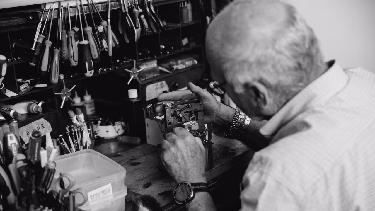 איש מבוגר מסבּר ומשפץ מנגנון מכני על שולחן עבודה עם כלי עבודה רבים תלויים ומונחים סביבו.