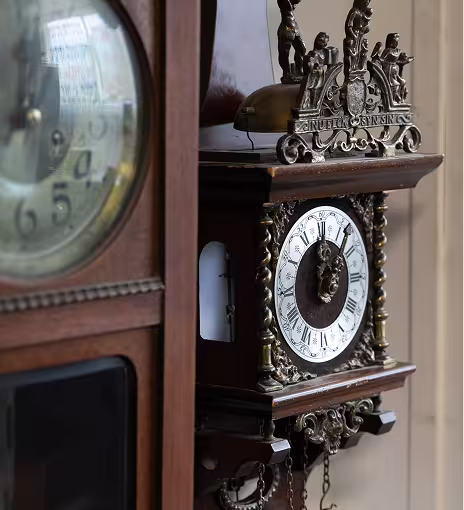 שעון קיר עתיק מעץ עם פנים עגול, ספרות רומיות וקישוטים מתכתיים מעוטרים.