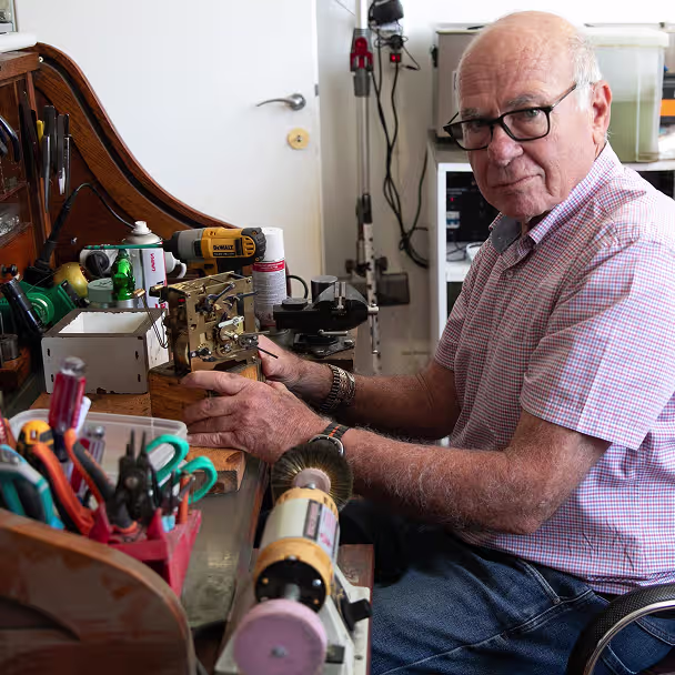 גבר מבוגר עם משקפיים יושב בסביבה של שולחן עבודה מלא בכלים ומחזיק מכשיר מכאני קטן.